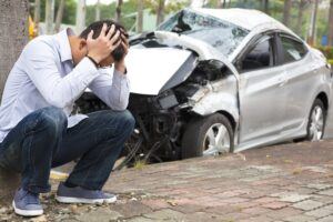 Worried driver sitting on the pavement next to damaged car from accident.